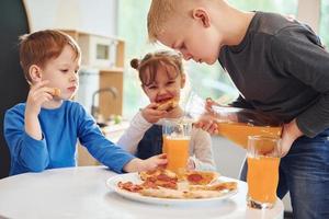 tres niños sentados en el interior junto a la mesa y comiendo pizza con jugo de naranja juntos foto