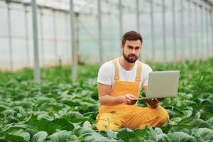 Young greenhouse worker in yellow uniform with laptop in hands have job inside of hothouse photo