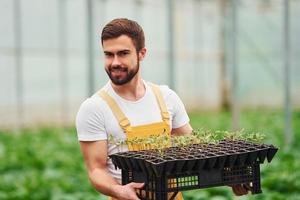 With black stand for plants in hands. Young greenhouse worker in yellow uniform have job inside of hothouse photo