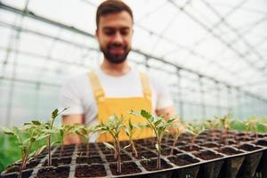 With black stand for plants in hands. Young greenhouse worker in yellow uniform have job inside of hothouse photo