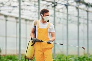 Young greenhouse worker in yellow uniform and white protective mask watering plants by using special equipment inside of hothouse photo