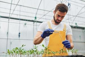 Holds test tube with water. Young greenhouse worker in yellow uniform have job inside of hothouse photo