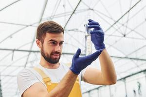 Holds test tube. Young greenhouse worker in yellow uniform have job inside of hothouse photo