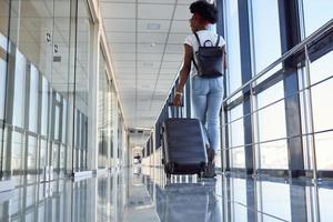 vista trasera si una joven pasajera afroamericana con ropa informal que está en el aeropuerto con equipaje foto