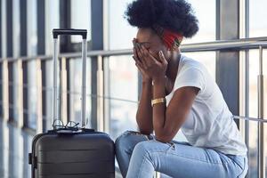 Sad and depressed young african american female passanger in casual clothes is in airport with baggage photo