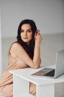 Beautiful brunette with curly hair in luxury clothes sits by table with laptop at home at daytime photo