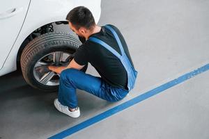 Man in work uniform sitting and changing car wheel indoors. Conception of automobile service photo