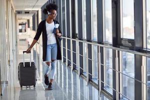 Young african american female passanger in casual clothes and headphones is in airport with baggage photo