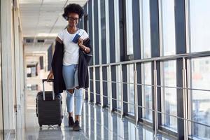 Young african american female passanger in casual clothes and headphones is in airport with baggage photo