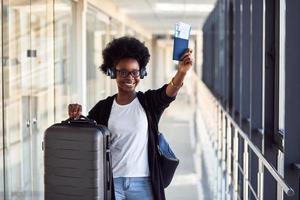 Young african american female passanger in casual clothes and headphones is in airport with baggage photo