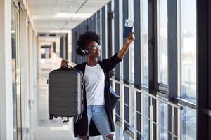 Young african american female passanger in casual clothes and headphones is in airport with baggage photo