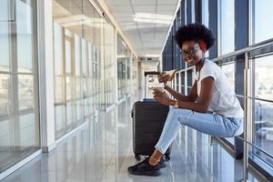 Young african american female passanger in casual clothes is in airport with baggage eating some food photo