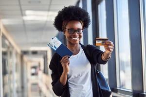 Young african american female passanger in casual clothes is in airport holding tickets and credit card photo