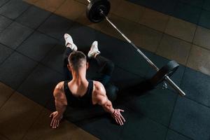 vista superior de un hombre fuerte con ropa deportiva que se sienta en el suelo y toma un descanso en el gimnasio foto