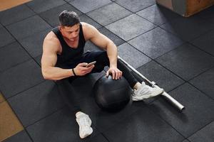 Strong man in sportive clothes sitting on the floor with phone in hands in the gym photo