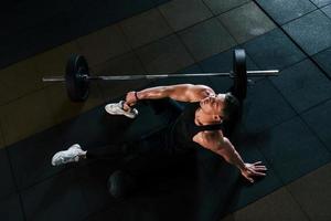 vista superior de un hombre fuerte con ropa deportiva que se sienta en el suelo y toma un descanso en el gimnasio foto