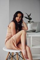 Beautiful brunette with curly hair in luxury clothes sits by table with laptop at home at daytime photo