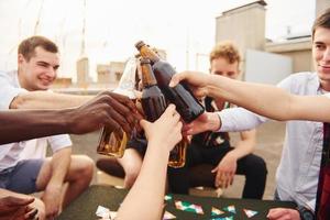 haciendo vítores por botellas con cerveza. un grupo de jóvenes con ropa informal tienen una fiesta en la azotea juntos durante el día foto