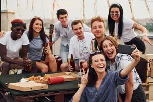 chica haciendo selfie. con deliciosa pizza. un grupo de jóvenes con ropa informal tienen una fiesta en la azotea juntos durante el día foto