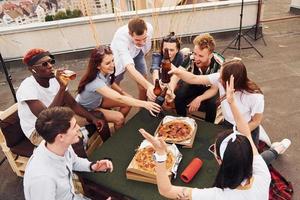 Doing cheers by bottles with beer. Group of young people in casual clothes have a party at rooftop together at daytime photo