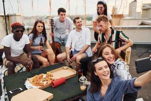 chica haciendo selfie. con deliciosa pizza. un grupo de jóvenes con ropa informal tienen una fiesta en la azotea juntos durante el día foto