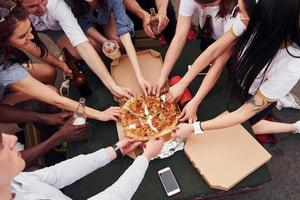 con deliciosa pizza. un grupo de jóvenes con ropa informal tienen una fiesta en la azotea juntos durante el día foto