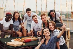 chica haciendo selfie. con deliciosa pizza. un grupo de jóvenes con ropa informal tienen una fiesta en la azotea juntos durante el día foto