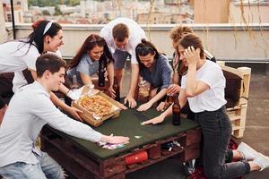 con deliciosa pizza. un grupo de jóvenes con ropa informal tienen una fiesta en la azotea juntos durante el día foto