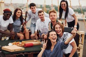 chica haciendo selfie. con deliciosa pizza. un grupo de jóvenes con ropa informal tienen una fiesta en la azotea juntos durante el día foto