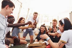 haciendo vítores por botellas con cerveza. un grupo de jóvenes con ropa informal tienen una fiesta en la azotea juntos durante el día