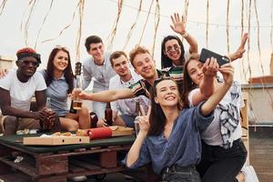 Girl making selfie. With delicious pizza. Group of young people in casual clothes have a party at rooftop together at daytime photo