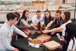Doing cheers by bottles with beer. Group of young people in casual clothes have a party at rooftop together at daytime photo