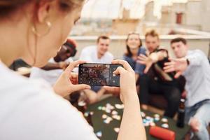 Girl doing photo when people playing card game. Group of young people in casual clothes have a party at rooftop together at daytime