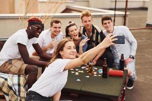 chica haciendo fotos cuando la gente juega a las cartas. un grupo de jóvenes con ropa informal tienen una fiesta en la azotea juntos durante el día