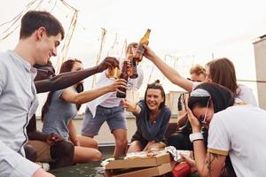 Doing cheers by bottles with beer. Group of young people in casual clothes have a party at rooftop together at daytime photo