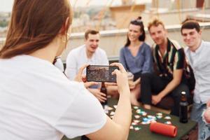 Girl doing photo when people playing card game. Group of young people in casual clothes have a party at rooftop together at daytime