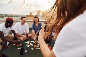 chica haciendo fotos cuando la gente juega a las cartas. un grupo de jóvenes con ropa informal tienen una fiesta en la azotea juntos durante el día
