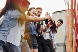 apoyándose en el borde de la azotea con decoraciones. grupo de jóvenes con ropa informal tienen una fiesta juntos durante el día foto