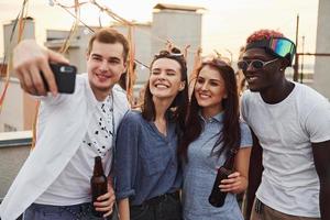 grupo de jóvenes con ropa informal tienen una fiesta en la azotea juntos durante el día y hacen selfie por teléfono