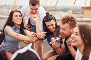 con deliciosa pizza. un grupo de jóvenes con ropa informal tienen una fiesta en la azotea juntos durante el día foto