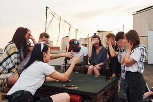 Sitting by table and playing card game. Group of young people in casual clothes have a party at rooftop together at daytime photo