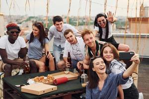 chica haciendo selfie. con deliciosa pizza. un grupo de jóvenes con ropa informal tienen una fiesta en la azotea juntos durante el día foto