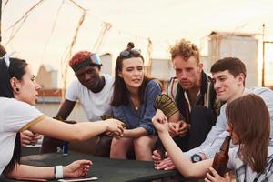 sentado junto a la mesa y jugando a las cartas. un grupo de jóvenes con ropa informal tienen una fiesta en la azotea juntos durante el día foto