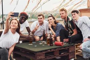 Playing game and sitting by the table. Group of young people in casual clothes have a party at rooftop together at daytime photo