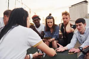 sentado junto a la mesa y jugando a las cartas. un grupo de jóvenes con ropa informal tienen una fiesta en la azotea juntos durante el día foto