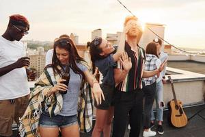 Playing game. Man's eyes covered by hands. Group of young people in casual clothes have a party at rooftop together at daytime photo