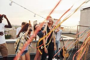 Playing game. Man's eyes covered by hands. Group of young people in casual clothes have a party at rooftop together at daytime photo