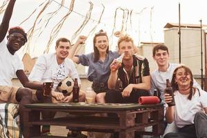 jugando y sentado junto a la mesa. un grupo de jóvenes con ropa informal tienen una fiesta en la azotea juntos durante el día foto