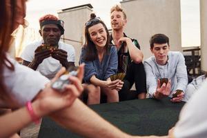 Sitting by table and playing card game. Group of young people in casual clothes have a party at rooftop together at daytime photo