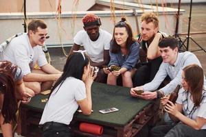 Sitting by table and playing card game. Group of young people in casual clothes have a party at rooftop together at daytime photo